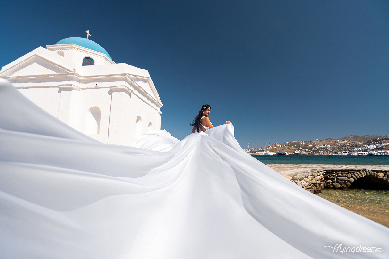 A woman in a flowing white dress poses near a white building with a blue dome by the sea in Mykonos, with her dress billowing dramatically in the wind against a clear blue sky.